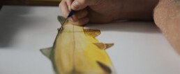A closeup of a man using a fine brush to refine a line on his painting of a gold and green fish.